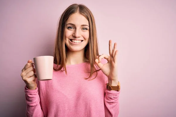 若いです美しいです赤毛女性飲むコーヒーのマグカップ以上隔離されたピンクの背景を行いますOkサインとともに指 優れたシンボル — ストック写真