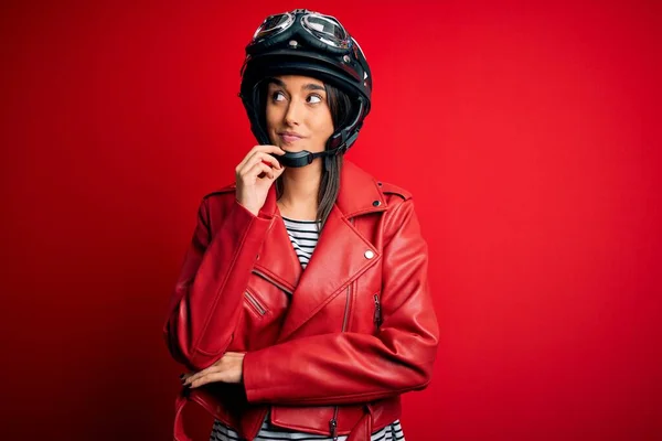 年轻美丽的黑发摩托车手 戴着摩托车头盔 穿着红色夹克 手放在下巴上思考着问题 沉思着表情 脸上挂着沉思的笑容 怀疑的概念 — 图库照片
