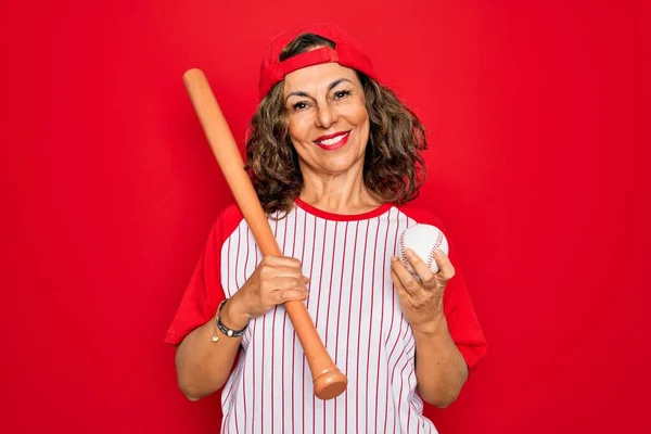 野球用具を身に着けている中年のシニア女性 幸せな顔立ちと歯を示す自信を持って笑顔で笑顔で赤い隔離された背景の上にバットとボール — ストック写真