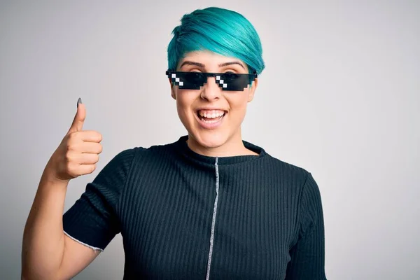 Young woman with blue fashion hair wearing thug life sunglasses over white background doing happy thumbs up gesture with hand. Approving expression looking at the camera showing success.