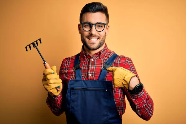 Young gardener man wearing working apron using gloves and tool over yellow background with surprise face pointing finger to himself