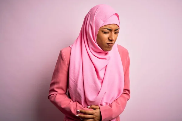 Young African American afro woman wearing muslim hijab over isolated pink background with hand on stomach because indigestion, painful illness feeling unwell. Ache concept.