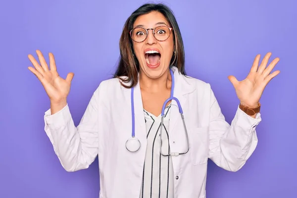 紫色の背景に聴診器と医療用コートを身に着けているプロの医師の女性は狂気を祝い 腕を上げ 興奮して叫んで目を開けて成功に驚いています 勝者のコンセプト — ストック写真