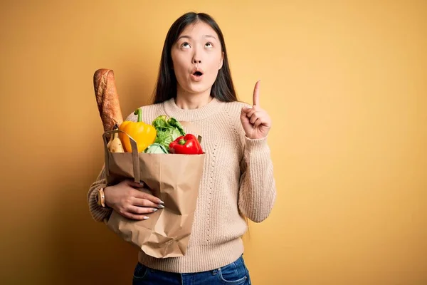 年轻的亚洲女人拿着一纸袋新鲜健康的食品放在黄色孤立的背景上 惊讶地抬起头 用手指和胳膊指指着 — 图库照片