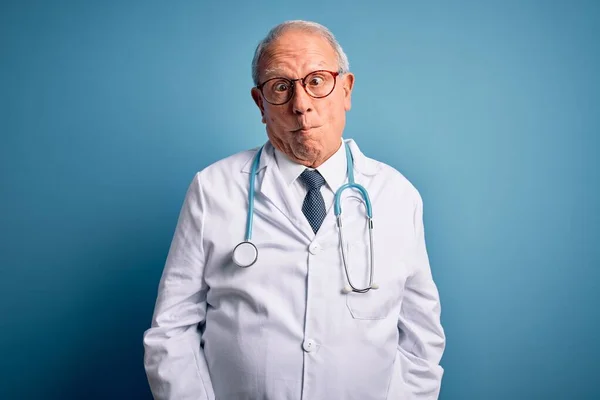 Senior grey haired doctor man wearing stethoscope and medical coat over blue background making fish face with lips, crazy and comical gesture. Funny expression.