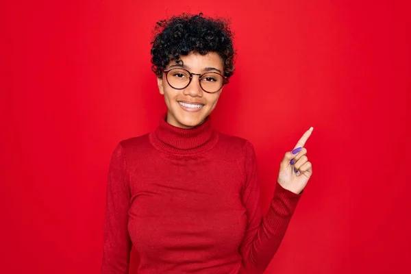 Young beautiful african american afro woman wearing turtleneck sweater and glasses with a big smile on face, pointing with hand finger to the side looking at the camera.