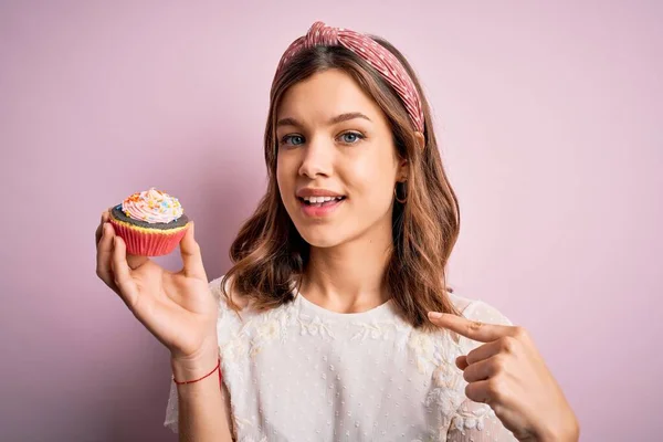 若いですブロンド女の子食べます甘いですチョコレートカップケーキ以上ピンク隔離された背景とともに驚き顔指して指へ彼自身 — ストック写真