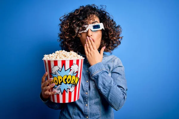 Young beautiful curly arab woman watching movie using 3d glasses eating popcorn snack cover mouth with hand shocked with shame for mistake, expression of fear, scared in silence, secret concept