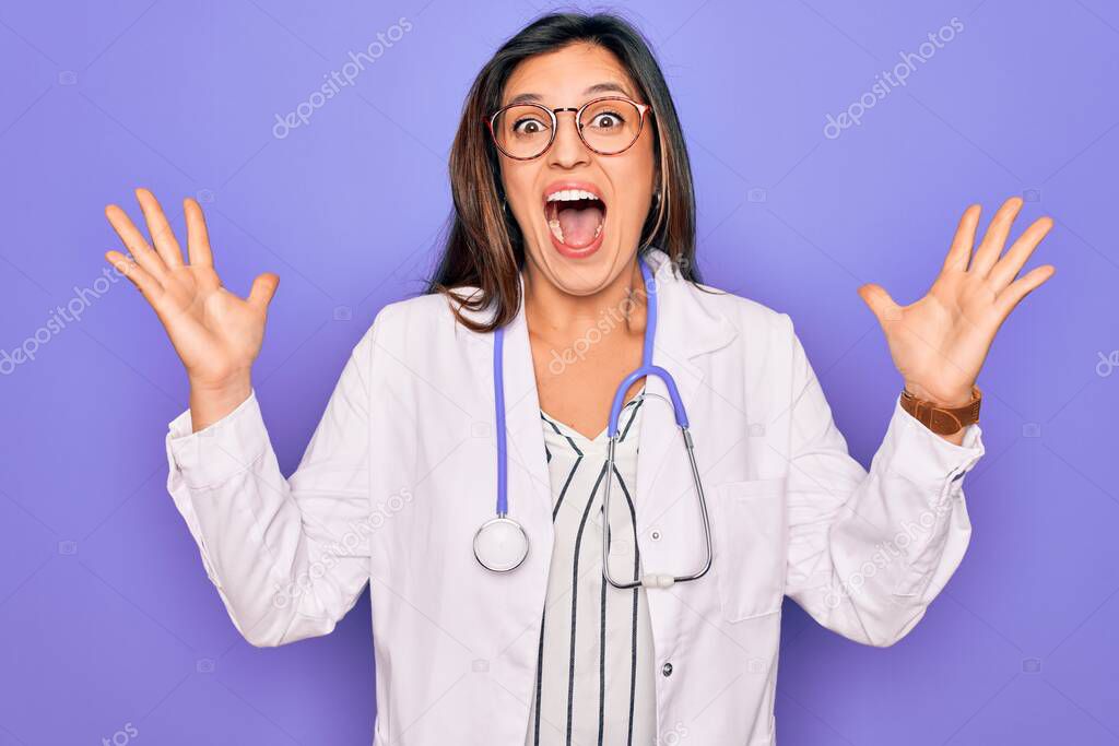Professional doctor woman wearing stethoscope and medical coat over purple background celebrating crazy and amazed for success with arms raised and open eyes screaming excited. Winner concept