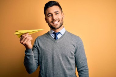 Elinde kağıttan bir uçak tutan genç, mutlu bir yüzü olan, kendine güvenen, gülümseyen ve dişlerini gösteren sarı arka planda duran yakışıklı bir adam.