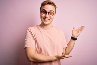 Genç, kızıl saçlı, günlük tişört giyen, soyutlanmış pembe arka planda duran genç adam gülümseyerek içeri girmeye davet ediyor.