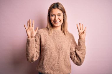 Pembe izole edilmiş arka plan üzerine kışlık yün kazak giyen genç ve güzel sarışın kadın dokuz numaralı parmaklarıyla işaret ederken kendinden emin ve mutlu bir şekilde gülümsüyor..