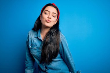 Genç esmer kadın, mavi izole edilmiş arka planda sıradan bir kot gömlek giyiyor kameraya bakıyor ve güzel ve seksi bir şekilde havada öpüşüyor. Aşk ifadesi.