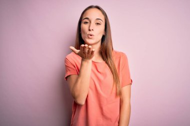 Genç, güzel bir kadın, rahat bir tişört giyip pembe arka planda dikiliyor ve kameraya bakıp havada öpücükler savuruyor. Güzel ve seksi. Aşk ifadesi.