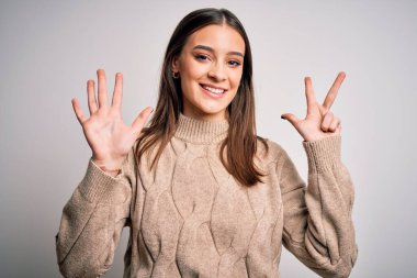 Genç, güzel, esmer, sıradan bir süveter giyen, beyaz arka plan üzerinde duran, 8 numaralı parmaklarıyla işaret eden, kendinden emin ve mutlu bir şekilde gülümseyen kadın..