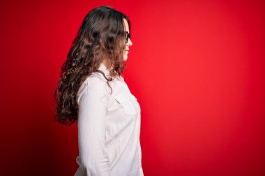 Kıvırcık saçlı, kırmızı arka planda gözlüklü ve tişörtlü, rahat bir görünüm takınan, kendine güveni olan güzel bir kadın..