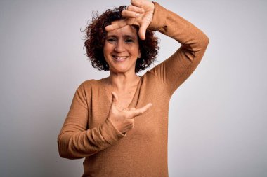 Orta yaşlı güzel kıvırcık saçlı kadın izole edilmiş beyaz arka plan üzerine günlük süveter giyip gülümsüyor ve elleri ve parmakları mutlu yüzlü bir çerçeve yapıyor. Yaratıcılık ve fotoğrafçılık kavramı.