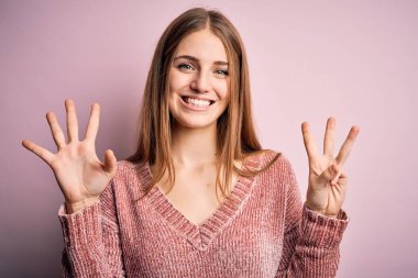 Genç, güzel, kızıl saçlı bir kadın pembe arka planda rahat bir süveter giyiyor. Sekiz numaralı parmaklarıyla işaret ediyor. Kendinden emin ve mutlu gülümsüyor..