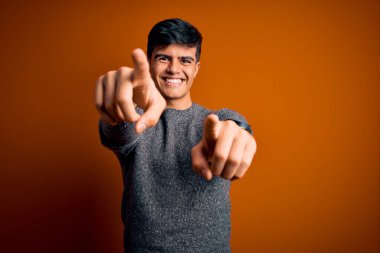 İzole edilmiş turuncu arka planda dikilen sıradan süveter giyen genç yakışıklı adam parmaklarıyla seni ve kamerayı işaret ediyor, neşeli ve pozitif gülümsüyor.