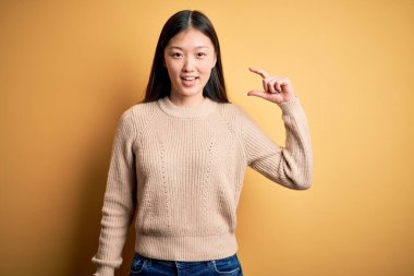 Sarı izole edilmiş arka plan üzerine günlük süveter giyen genç, güzel Asyalı kadın gülümseyen ve kendine güvenen el hareketleriyle küçük beden işareti yapan parmaklarıyla ve kamerasıyla. Ölçü kavramı.