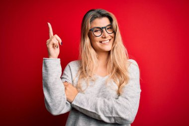 İzole edilmiş kırmızı arka planda süveter ve gözlük takan genç, sarışın bir kadın. Yüzünde kocaman bir gülümseme, eli ve parmağı kameraya bakıyor..