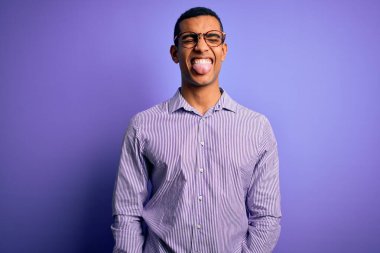 Çizgili gömlek ve mor arka planda gözlük takan yakışıklı Afro-Amerikan adam komik bir ifadeyle dilini dışarı çıkarıyor. Duygu kavramı.