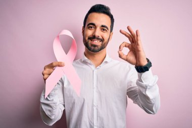 Arkaplanda pembe kanser kurdelesi tutan sakallı yakışıklı genç adam parmaklarıyla imza atıyor, mükemmel bir sembol.