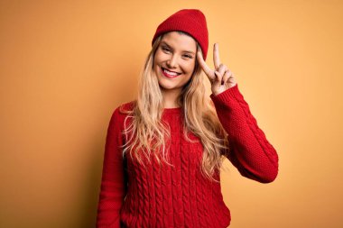 Genç, sarışın, rahat bir süveter giyen, beyaz arka planda yün şapkalı, iki numaralı parmaklarıyla iki numarayı gösteren, kendinden emin ve mutlu gülümseyen bir kadın..