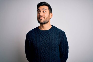 Sakallı, sıradan bir süveter giyen, beyaz arka planda durup gülümseyen ve kafasını çeviren genç yakışıklı bir adam..