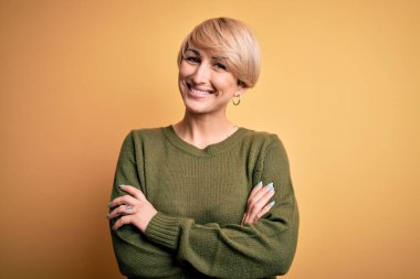 Modern kısa saçlı, sarı arka planda rahat süveter giyen, kollarını kavuşturup kameraya gülümseyen genç sarışın kadın. Pozitif kişi.