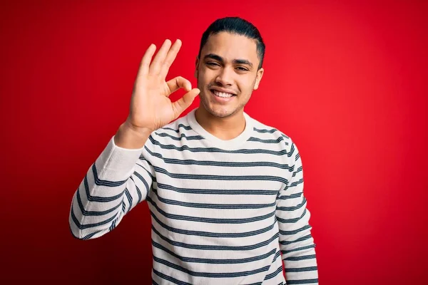 Young brazilian man wearing casual striped t-shirt standing over isolated red background smiling positive doing ok sign with hand and fingers. Successful expression.