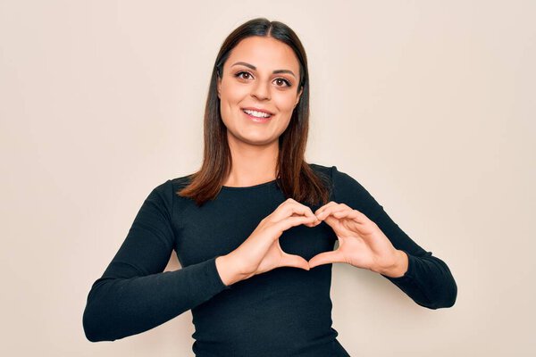 Молодая красивая брюнетка в повседневной футболке стоит на изолированном белом фоне улыбаясь в любви показывая символ сердца и форму с руками. Романтическая концепция.