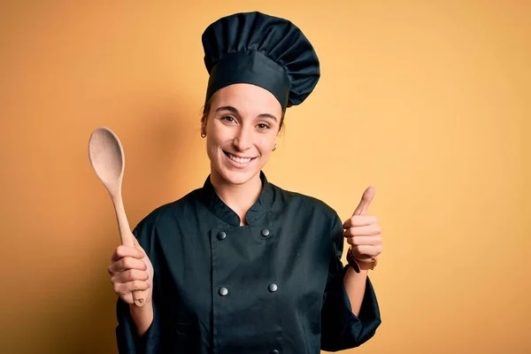 年轻美丽的厨师女士身穿炊具制服 头戴木勺 喜形于色 做手势 用手指竖起大拇指 是极好的标志 — 图库照片