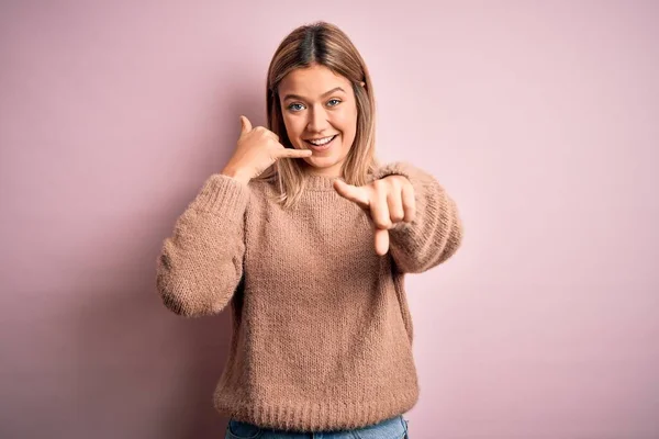 年轻美丽的金发女人 穿着冬季羊毛毛衣 背景是粉色的 背景是孤立的 微笑着在电话上说话 指着你 打给我 — 图库照片