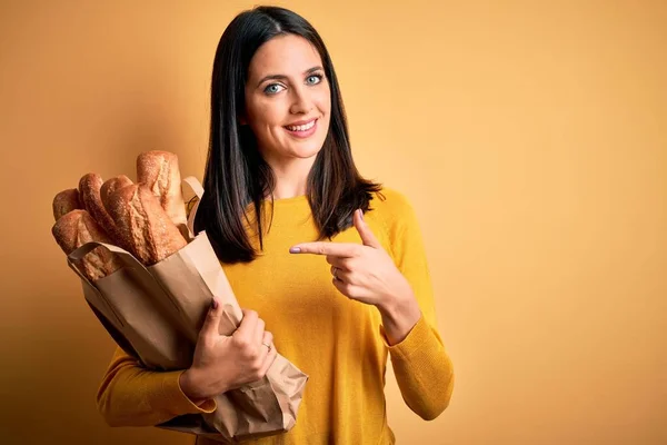 一个蓝眼睛的年轻女人拿着装有面包的纸袋 披着孤立的黄色背景 非常高兴地用手和手指指指点点 — 图库照片