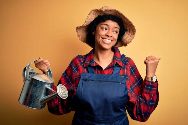 若いですアフリカ系アメリカ人の農家の女性との巻き毛を身に着けているエプロンと帽子を使用して水を指し 幸せな顔を笑顔で親指で側面まで示すことができます — ストック写真