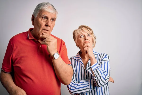 Senior beautiful couple standing together over isolated white background with hand on chin thinking about question, pensive expression. Smiling with thoughtful face. Doubt concept.