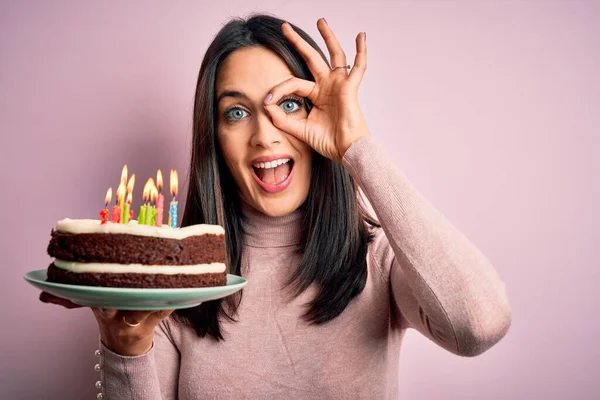 Young woman with blue eyes holding birthday cake with candles over pink background with happy face smiling doing ok sign with hand on eye looking through fingers