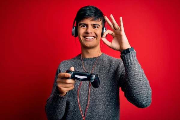 Young handsome gamer man playing video game using joystick and headphones doing ok sign with fingers, excellent symbol