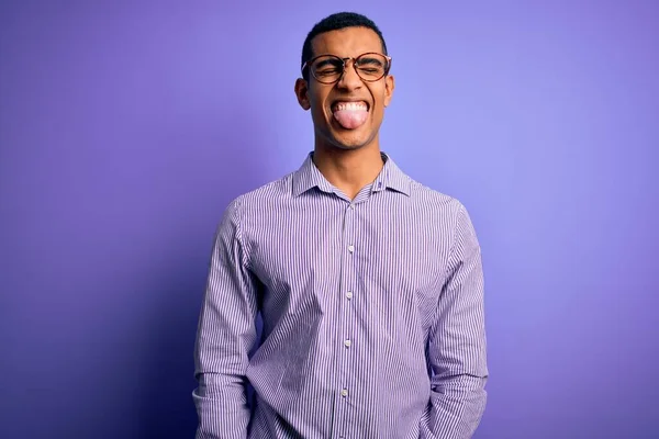 紫色の背景にストライプのシャツとメガネを身に着けているハンサムなアフリカ系アメリカ人男性が面白い表情で幸せ舌を突き出しています 感情概念 — ストック写真