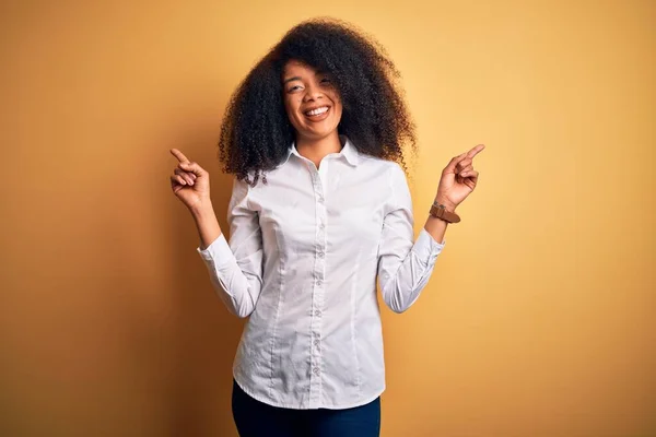 黄色の背景の上に立つアフロの髪を持つ若い美しいアフリカ系アメリカ人のエレガントな女性は 異なる方向への指で自信を持って指して笑顔 広告のコピースペース — ストック写真