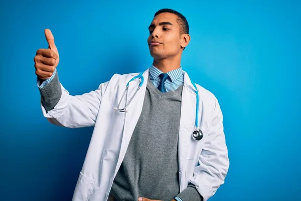 英俊的非洲裔美国医生 身穿外套 带着听诊器 背景是蓝色的 他看上去很自豪 微笑着向旁边竖起大拇指 — 图库照片