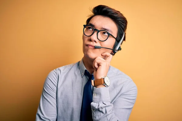 Young handsome chinese call center agent man wering glasses using headset with hand on chin thinking about question, pensive expression. Smiling with thoughtful face. Doubt concept.