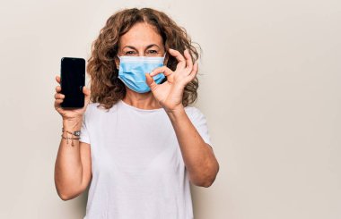 Koronavirüs maskesi takan orta yaşlı bir kadın akıllı telefon boş ekranını gösteriyor. Parmaklarıyla işaret veriyor, gülümsüyor.
