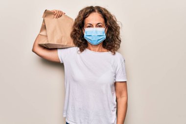 Orta yaşlı teslimatçı kadın Coronavirus koruma maskesi takıyor ve elinde kağıt torba var. Pozitif ve mutlu görünüyor. Kendine güvenen bir gülümsemeyle dişlerini gösteriyor.