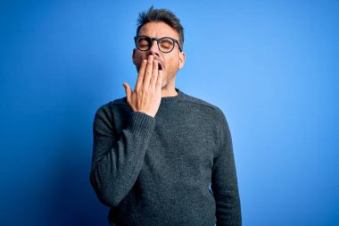 Mavi arka planda duran sıradan süveter ve gözlük takan genç yakışıklı adam ağzıyla esnemekten sıkılmış. Huzursuz ve uykulu..