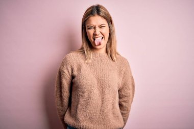 Pembe izole arka plan üzerine kışlık yün kazak giyen güzel sarışın kadın komik bir ifadeyle dilini dışarı çıkartıyor. Duygu kavramı.