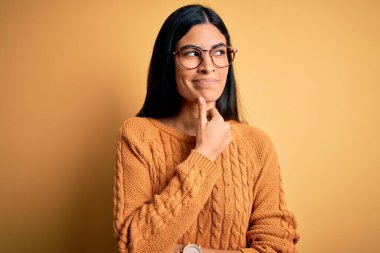 Sarı izole arka planda gözlük takan genç, İspanyol bir kadın. Elleri çenesinde soru sorarken, düşünceli bir ifade takınıyor. Düşünceli bir yüzle gülümsüyor. Şüphe kavramı.