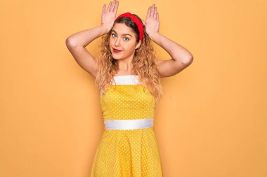 Sarı arka planda dikey duran mavi gözlü güzel sarışın kadın avuçlarında alaycı ve şüpheci ellerle tavşan kulağı hareketi yapıyor. Paskalya tavşanı kavramı.