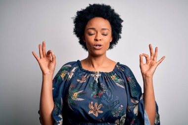 Kıvırcık saçlı, çiçekli bir elbise giyen, gözleri kapalı gülümseyen ve meditasyon yaparken parmaklarıyla gülümseyen güzel Afro-Amerikan kadın. Yoga konsepti.